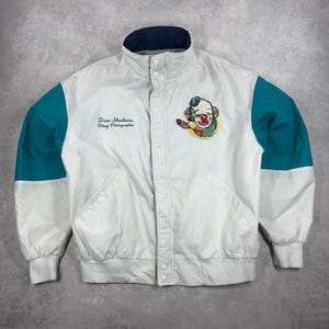 Puede incluir: Chaqueta blanca con mangas turquesas y cuello azul marino. La chaqueta presenta un parche de payaso colorido y el texto "Dane Sherburne, Chief Photographer". La chaqueta tiene cierre de botones y puños acanalados.