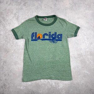 Puede incluir: Camiseta verde con cuello y mangas verde oscuro. La palabra "Florida" está impresa en azul en la parte delantera, con un sol amarillo y un gráfico de velero azul. La camiseta está hecha de un material suave y texturizado.
