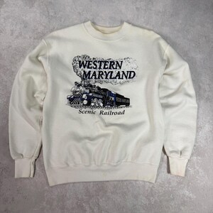 May include: Cream-colored sweatshirt featuring a graphic of a steam train with the text "WESTERN MARYLAND Scenic Railroad" in blue and black. The sweatshirt has long sleeves and ribbed cuffs.