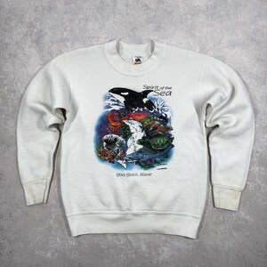May include: White sweatshirt with a graphic of marine life, including an orca, sea turtles, and fish. The text "Spirit of the Sea" and "York Beach, Maine" are printed on the front. The sweatshirt has a crew neck and long sleeves.