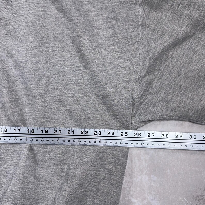 May include: A close-up of a gray t-shirt with a measuring tape across the bottom. The tape measures from 16 to 30 inches. The shirt is a solid color and appears to be made of a soft material.