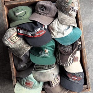 Op de afbeelding: Een houten kist vol met baseballpetten. De petten tonen logo's en merken van Harley-Davidson, John Deere, Coors Light en andere merken. Kleuren zijn onder meer groen, zwart, grijs en camouflagepatronen. De petten zijn gemaakt van katoen en andere stoffen.