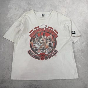 May include: White t-shirt featuring a graphic of the Chicago Bulls basketball team with the text "1991 NBA Champs, 1992, 1993, 1996". The shirt has a crew neck and short sleeves. A Starter logo is on the sleeve.
