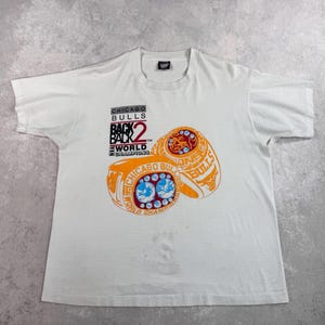 May include: White t-shirt featuring a graphic of two orange and blue championship rings. The text on the shirt reads "CHICAGO BULLS BACK 2 BACK WORLD CHAMPIONS."