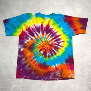 May include: A colorful tie-dye t-shirt with a spiral design. The shirt features a vibrant mix of colors, including blue, yellow, orange, purple, and red. The short-sleeved shirt is laid flat, showcasing the intricate dye pattern.