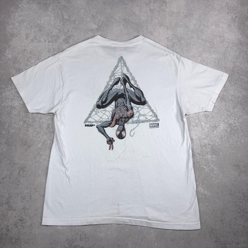 May include: White t-shirt featuring a graphic of Spider-Man hanging upside down within a triangular web design. The design includes the text "HUF" and "MARVEL".