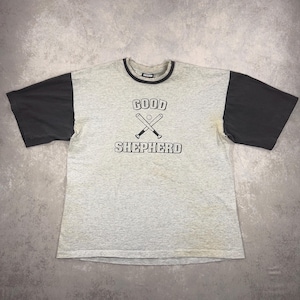 May include: A grey t-shirt with black sleeves. The front displays the text "GOOD SHEPHERD" above crossed baseball bats. The collar is black. The shirt has a vintage look.