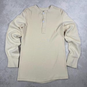 Könnte beinhalten: Cremefarbenes langärmliges Henley-Shirt mit gerippter Struktur. Das Shirt hat eine Knopfleiste mit drei Knöpfen und gerippte Bündchen. Der Stoff scheint ein weiches, bequemes Material zu sein.