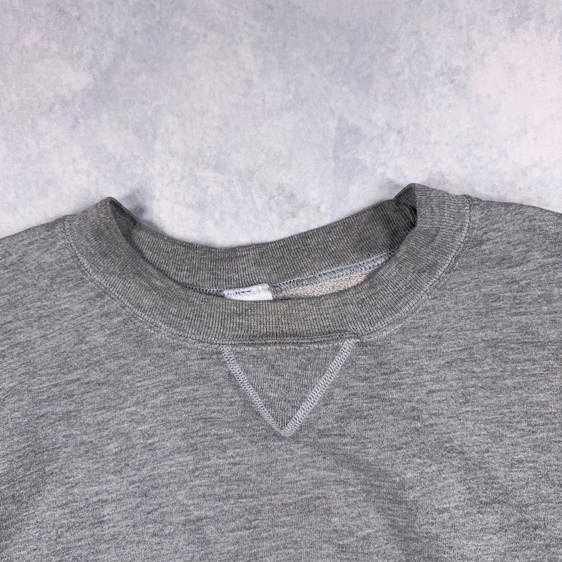 May include: Close-up of a heather gray sweatshirt. The crew neck collar and the V-shaped stitching at the neckline are visible. The fabric appears soft and comfortable, suitable for casual wear. The garment is a solid color.