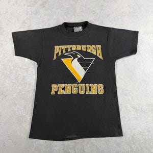 Könnte beinhalten: Schwarzes T-Shirt mit dem Logo der Pittsburgh Penguins in Gelb und Weiß. Das Logo zeigt einen Pinguin in einem gelben Dreieck, mit dem Teamnamen darüber und darunter.