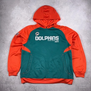 Puede incluir: Una sudadera con capucha naranja y verde azulado de los Dolphins con una capucha y un bolsillo delantero. La palabra "DOLPHINS" está impresa en blanco en el pecho, con el logotipo del equipo encima. Las mangas y la capucha son naranjas, mientras que el cuerpo es verde azulado.