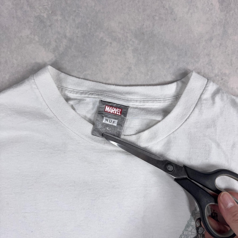 May include: A white t-shirt with a crew neck and the Marvel logo on the tag. The tag also has the Huf logo and the size L. A pair of scissors is cutting the tag.