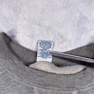 May include: Close-up of a gray sweatshirt collar with a white tag. The tag includes washing instructions and fabric content: 53% polyester, 47% cotton. The tag also includes the manufacturer's information.