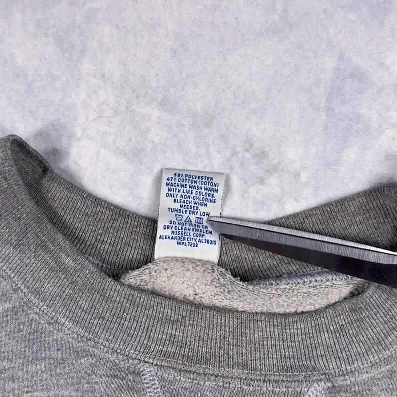 May include: Close-up of a gray sweatshirt collar with a white tag. The tag includes washing instructions and fabric content: 53% polyester, 47% cotton. The tag also includes the manufacturer's information.