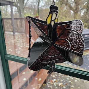 May include: A stained glass suncatcher in the shape of a figure with outstretched arms, featuring brown, black, yellow, and white glass pieces. A beaded strand hangs from the figure's hand. The suncatcher is suspended in front of a window.