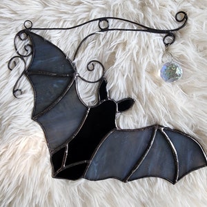 May include: A stained glass bat with a black body and gray wings hanging from a metal wire with a crystal prism hanging from the end.