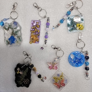 May include: A collection of handmade keychains with various designs. Keychains feature resin pendants with dried flowers, butterflies, and bee motifs. Each keychain has a metal clasp and decorative beads. The color palette includes blues, purples, yellows, and pinks.