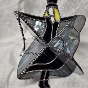 May include: A stained glass suncatcher in the shape of a figure with outstretched arms. The figure is primarily black and gray, with yellow and white accents. A beaded chain hangs from the top. The suncatcher is designed to hang in a window.