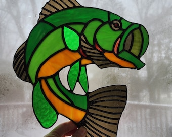 May include: Stained glass fish in shades of green, orange, and brown. The fish has a detailed design with black outlines, and is held up against a window. The fish is a decorative item, possibly for a window or wall.
