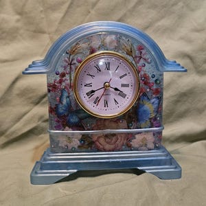 May include: A mantel clock with a light blue arched frame containing a round clock face with black Roman numerals and quartz movement. The interior is filled with pressed pink and white flowers, small pearls, and two blue butterfly images.