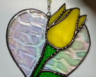 May include: Stained glass heart-shaped suncatcher with a yellow tulip and iridescent glass. The tulip has green leaves and stem. The suncatcher is trimmed in a dark metal and has a beaded hanging chain.
