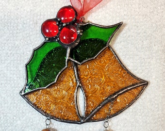 Puede incluir: Un adorno navideño de vidrio emplomado con forma de campana con una rama de acebo y bayas rojas en la parte superior. La campana es dorada con una superficie texturizada y tiene dos cuentas colgantes con diseños florales.