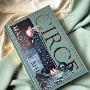 May include: A teal blue hardcover book with gold lettering titled "Circe" by Madeline Miller. The cover features an illustration of a woman pouring water from a pitcher.