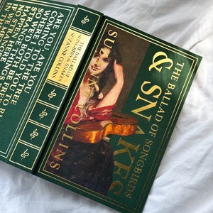 May include: A green hardcover book with gold lettering titled "The Ballad of Songbirds & Snakes" by Suzanne Collins. The cover features a painting of a woman playing a guitar.