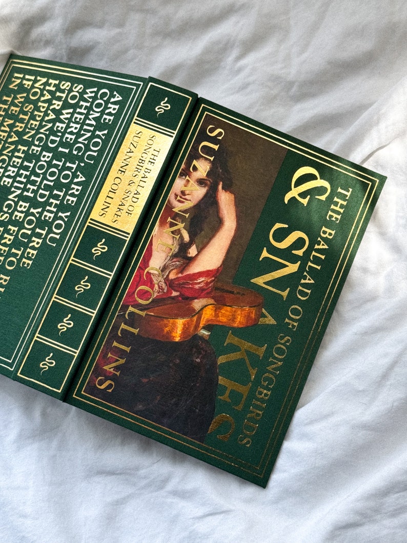May include: A green hardcover book with gold lettering titled "The Ballad of Songbirds & Snakes" by Suzanne Collins. The cover features a painting of a woman playing a guitar.