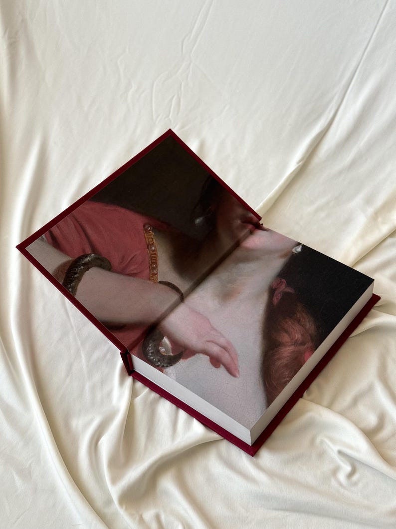 May include: A burgundy hardcover book with a painting of a woman's arm and shoulder on the cover. The woman is wearing a red dress and a gold bracelet.