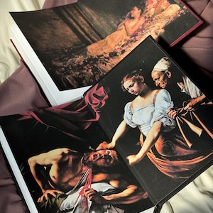 Puede incluir: Dos libros abiertos con cubiertas negras y p&aacute;ginas blancas. El libro superior presenta una pintura de una mujer acostada en una cama de flores. El libro inferior presenta una pintura de una mujer con un vestido blanco sosteniendo la cabeza de un hombre en sus manos. El hombre est&aacute; acostado en una s&aacute;bana blanca con una cuerda roja alrededor del cuello.
