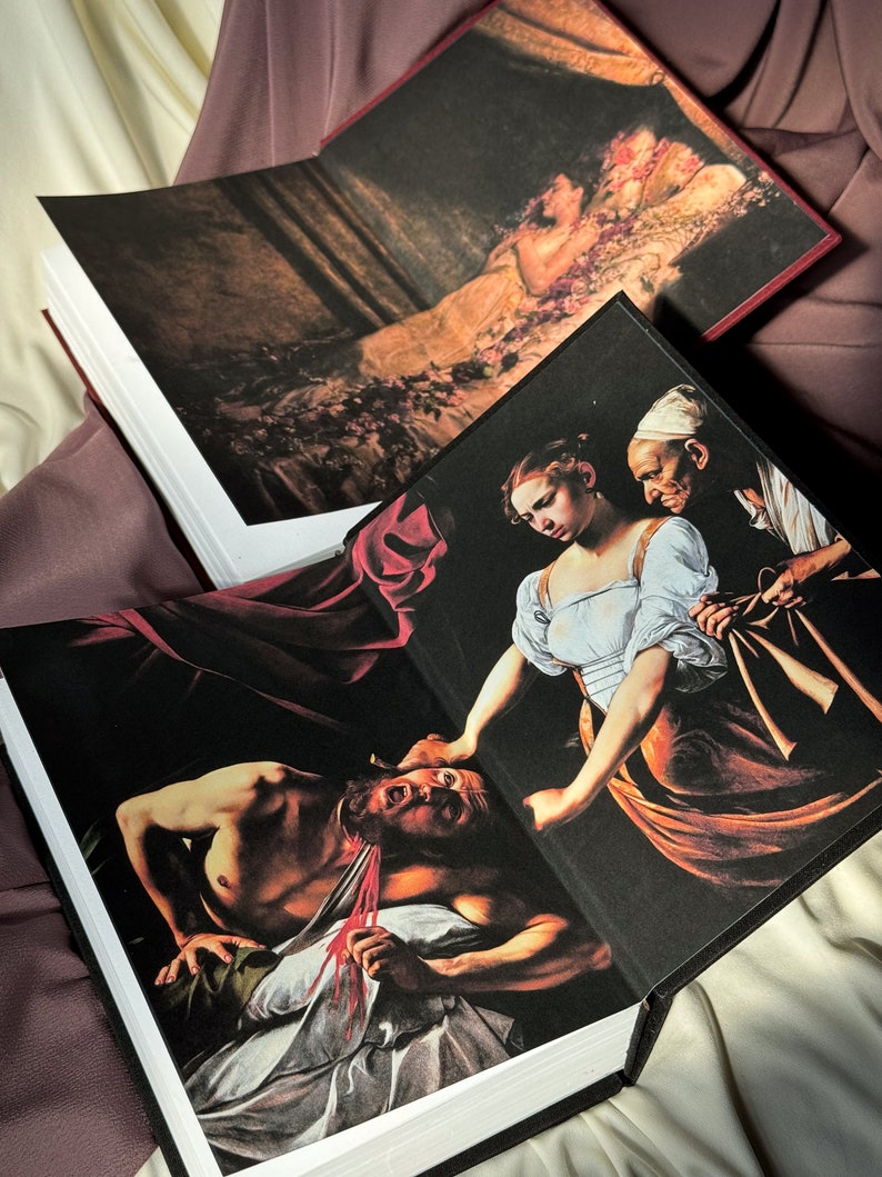 Puede incluir: Dos libros abiertos con cubiertas negras y p&aacute;ginas blancas. El libro superior presenta una pintura de una mujer acostada en una cama de flores. El libro inferior presenta una pintura de una mujer con un vestido blanco sosteniendo la cabeza de un hombre en sus manos. El hombre est&aacute; acostado en una s&aacute;bana blanca con una cuerda roja alrededor del cuello.