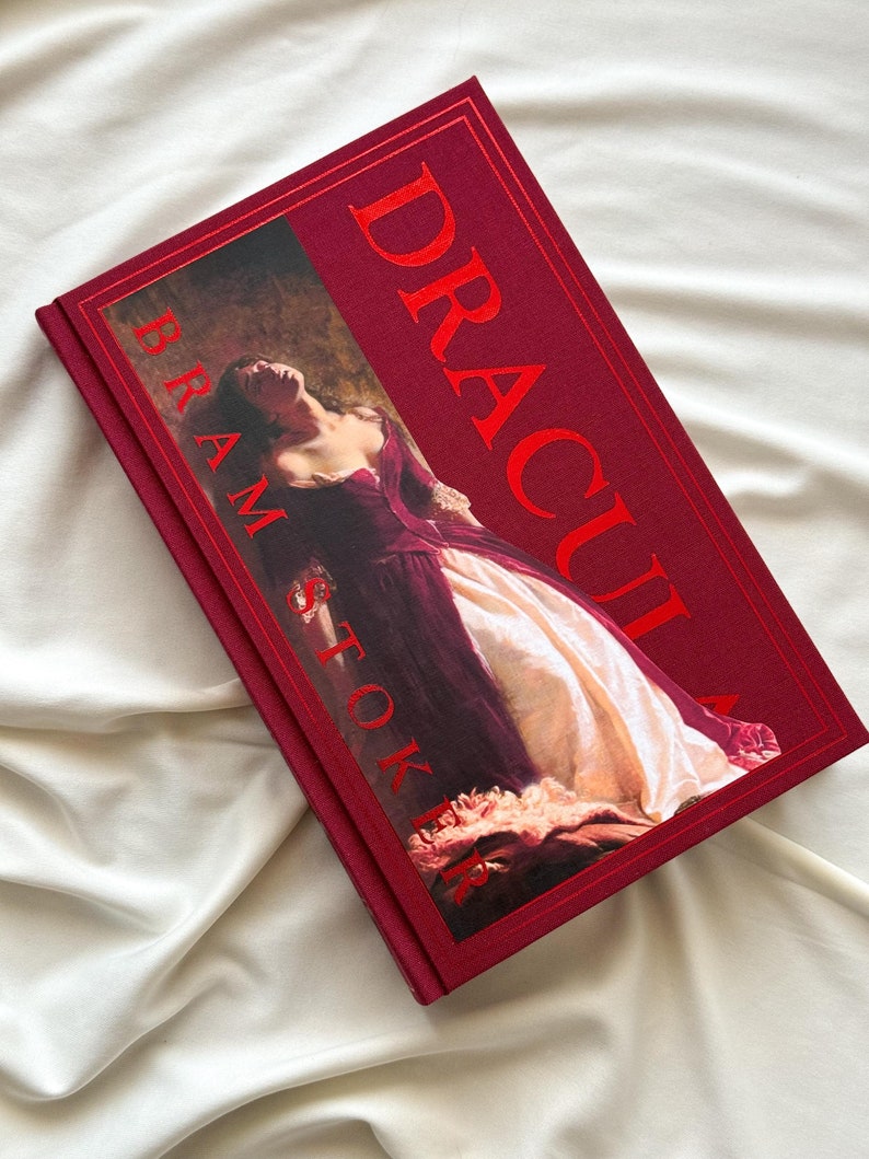 May include: A red hardcover book with the title "Dracula" in gold lettering on the spine. The front cover features a painting of a woman in a white and purple dress.