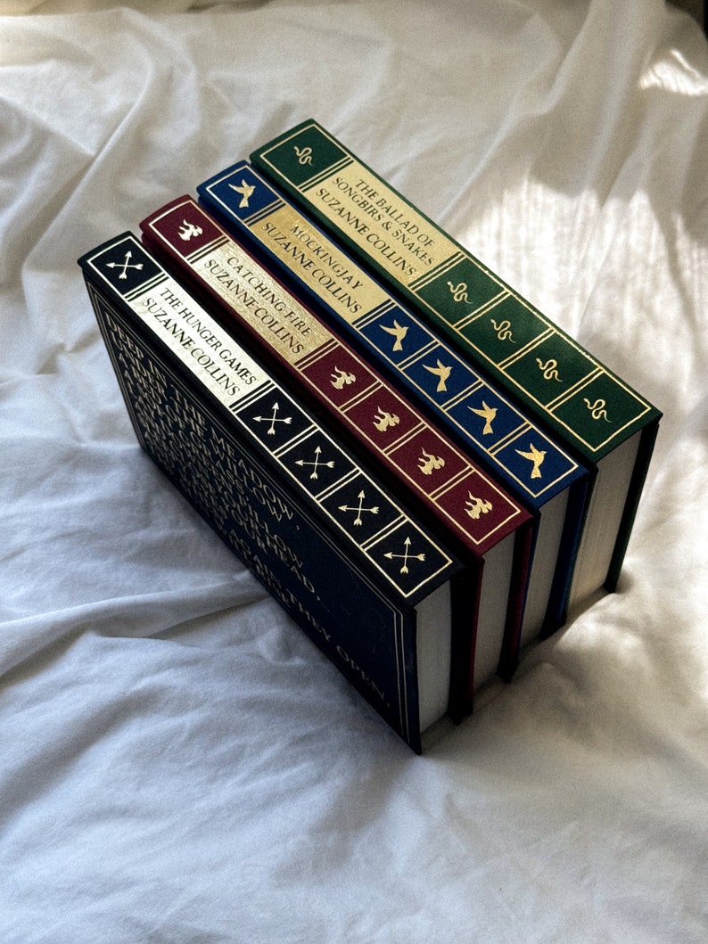 May include: A stack of five hardcover books with gold lettering and illustrations on the spines. The titles are "The Hunger Games", "Catching Fire", "Mockingjay", "The Ballad of Songbirds and Snakes", and "The Hunger Games".
