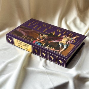 May include: A purple hardback book with gold lettering titled "If We Were Villains" by M.L. Rio. The cover features a painting of a group of people in a dramatic scene.