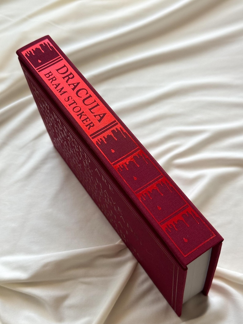 May include: A burgundy hardcover book with the title "Dracula" by Bram Stoker printed in red on the spine. The spine is decorated with dripping blood-like red accents.