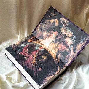 May include: An open book with a purple cover and a painting of a scene from the Bible. The painting depicts a group of people in a dark, shadowy setting. The book is lying open on a white fabric surface.