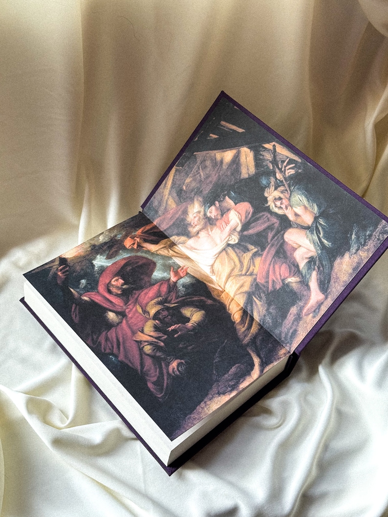May include: An open book with a purple cover and a painting of a scene from the Bible. The painting depicts a group of people in a dark, shadowy setting. The book is lying open on a white fabric surface.