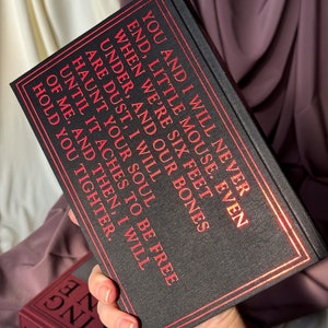 Puede incluir: Un libro de tapa dura negro con letras doradas. El texto del libro dice: "You and I will never end, little mouse. Even when we're six feet under, and our bones are dust, I will haunt your soul until it aches to be free of me. And then, I will hold you tighter."