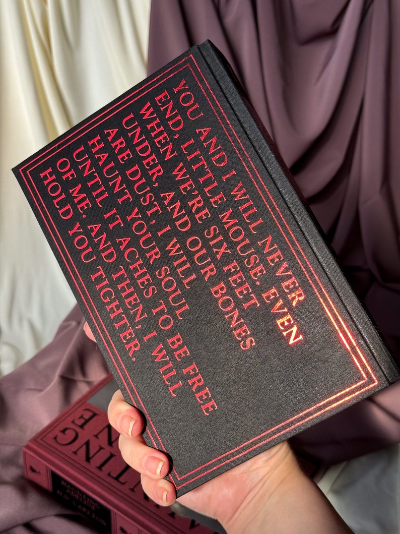 Puede incluir: Un libro de tapa dura negro con letras doradas. El texto del libro dice: "You and I will never end, little mouse. Even when we're six feet under, and our bones are dust, I will haunt your soul until it aches to be free of me. And then, I will hold you tighter."