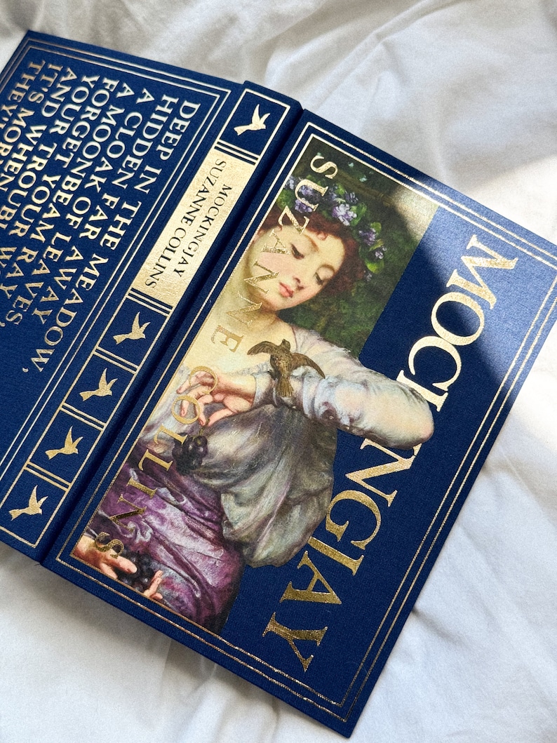 May include: A blue hardcover book with gold lettering titled "Mockingjay" by Suzanne Collins. The cover features a painting of a woman with a bird in her hand.