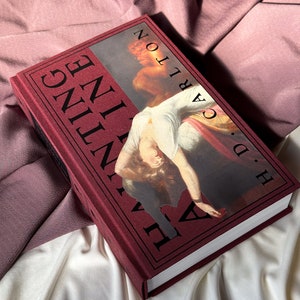 Puede incluir: Un libro de tapa dura color burdeos con el t&iacute;tulo "The Vanishing Half" de Brit Bennett. La portada presenta una pintura de una mujer con un vestido blanco.