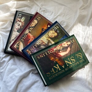 May include: Four hardcover books stacked on top of each other. The books are all green, blue, red, and black with gold lettering. The titles of the books are "The Ballad of Songbirds & Snakes", "Suzanne Collins", "Suzanne Collins", "Suzanne Collins", and "Suzanne Collins".