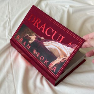 May include: A red hardcover book titled "Dracula" by Bram Stoker. The cover features a painting of a woman lying down in a white dress.