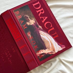May include: A red hardcover book with the title "Dracula" and the author "Bram Stoker" printed in gold lettering. The cover features a painting of a woman in a white dress.