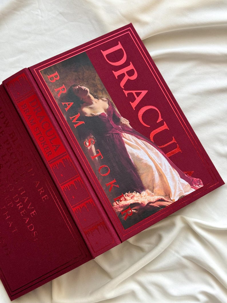 May include: A red hardcover book with the title "Dracula" and the author "Bram Stoker" printed in gold lettering. The cover features a painting of a woman in a white dress.