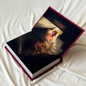 May include: A burgundy hardcover book with a painting of a woman lying in bed on the front cover. The book is open to reveal a similar painting on the inside pages.