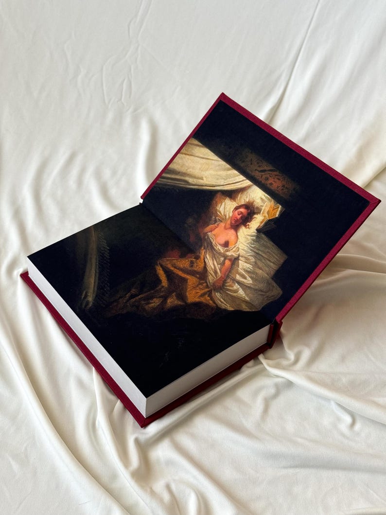 May include: A burgundy hardcover book with a painting of a woman lying in bed on the front cover. The book is open to reveal a similar painting on the inside pages.