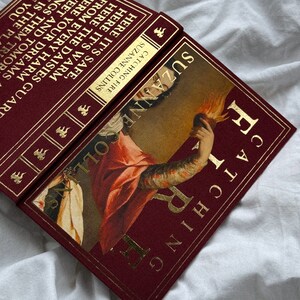 May include: A burgundy hardback book with gold lettering and a gold-trimmed spine. The book title is "Catching Fire" by Suzanne Collins. The cover features a painting of a woman holding a flaming heart.