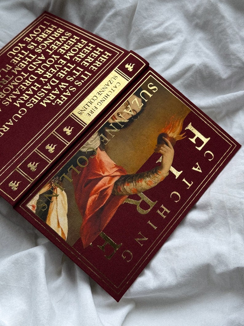 May include: A burgundy hardback book with gold lettering and a gold-trimmed spine. The book title is "Catching Fire" by Suzanne Collins. The cover features a painting of a woman holding a flaming heart.